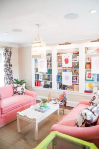 Beautiful historic home library located on the landing of the stairway, featuring a pair of pink sofas around a white lacquer coffee table. Library of colorful books with custom painted silhouettes by Carter Kustera. Modern family living in an historic neighborhood in Oklahoma City www.pencilshavingsstudio.com